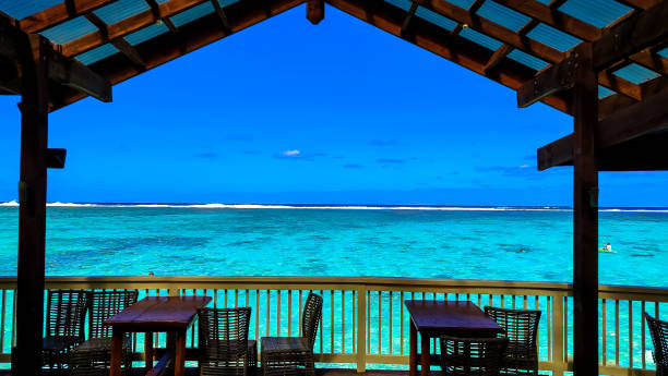 istock Tranquil lagoon stretches out from covered deck, Cook Islands. Crystal-clear water laps at pristine white sand, distant surfers ride playful waves. Perfect island getaway awaits. 2178797550