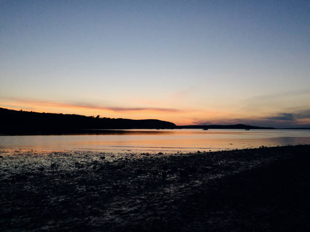 A breathtaking image of the sun setting over a tranquil beach in Maine. The sky is filled with a blend of blue, orange, black, and golden hues, which beautifully reflect off the calm ocean water. The contrast between the vivid colors of the sunset and the darkening beach creates a serene and peaceful atmosphere, capturing the beauty of nature as day turns to night.