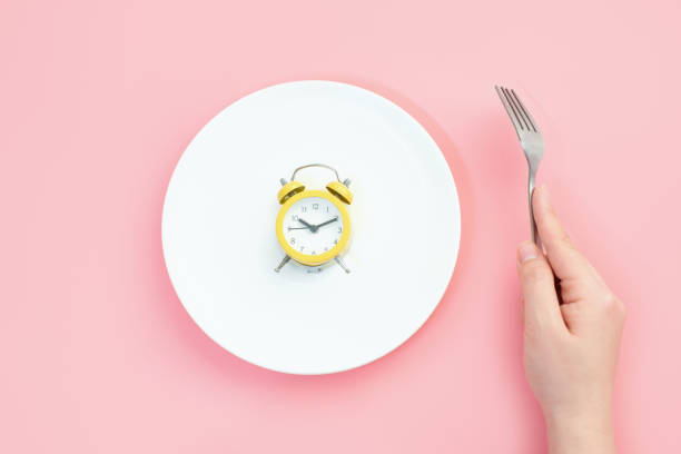 istock Female hand holding a fork next to an empty plate with alarm clock on a pink background close-up top view. 2178812162