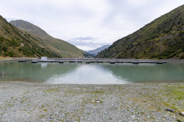 A row of solar panels installed near a calm reservoir in a mountainous region. The panels are set against a backdrop of rugged hills.