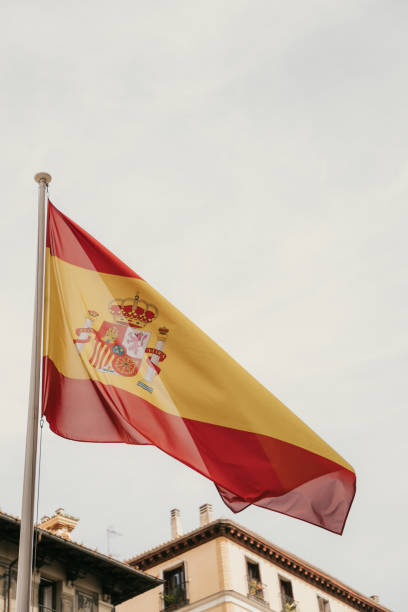 A gentle breeze catches the vibrant red and yellow of the Spanish flag, waving proudly amidst the classic architecture of Madrid. The scene is framed by charming buildings, their balconies adorned with wrought iron details that evoke the city’s rich history. A nearby vintage lamppost adds a touch of old-world charm, contrasting beautifully with the timeless patriotism symbolized by the flag. This image captures the essence of Madrid’s identity, where heritage and national pride blend seamlessly into the rhythm of urban life.