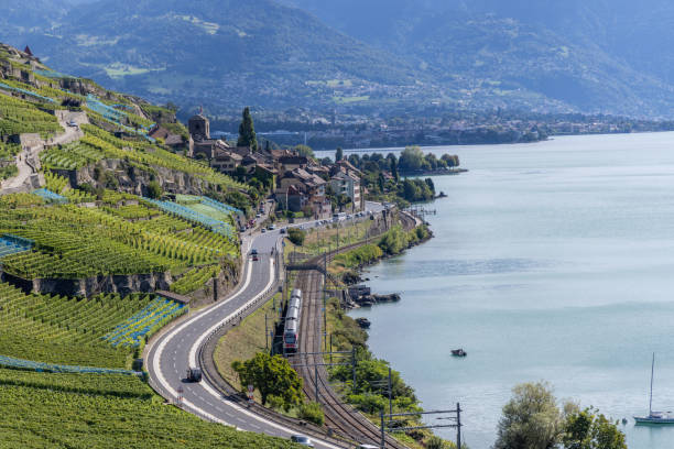 A scenic lakeside landscape featuring terraced vineyards, a winding road, and a train track along the shoreline, with a picturesque village nestled by the water and mountains in the background.