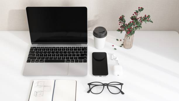 Desktop of modern white desk with laptop computer, smartphone, earphone, notebook and eyeglasses in sunlight from window on beige wall for home office working, product background 3D