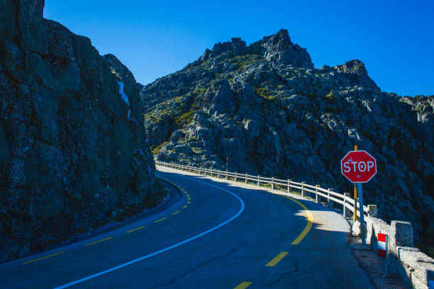 A curving mountain road bordered by rocky cliffs and a clear blue sky, with a stop sign positioned along the roadside, creating a dramatic and adventurous atmosphere