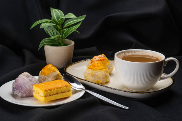 Closeup view of hot milk coffee in white ceramic cup with chinese pastry moon cake in white ceramic dark edge tray, colorful chinese pastry moon cake and layer cake in white ceramic dish on black background.