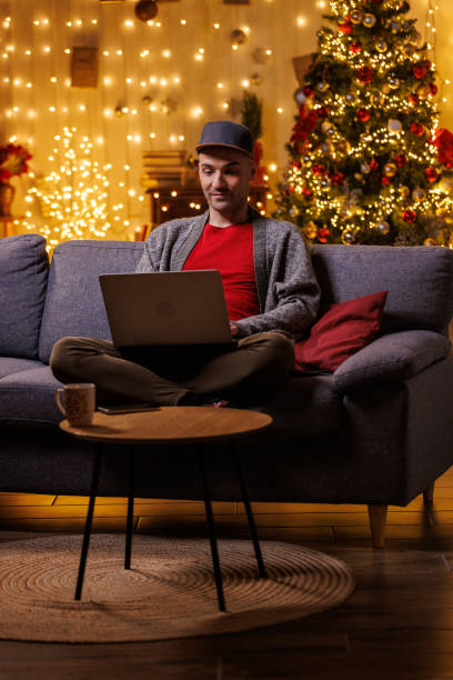 istock Young man, using laptop, sitting on the sofa with Christmas tree in the background 2178866271