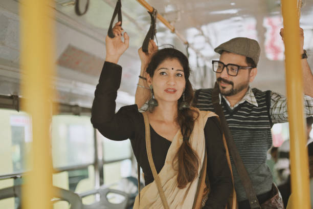 A mid-adult couple stands side by side on a public bus, sharing a quiet moment together. Holding onto the overhead rail and a nearby pole for balance, they exchange few words as they travel.
