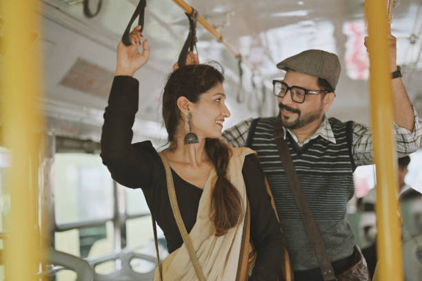 A mid-adult couple stands side by side on a public bus, sharing a quiet moment together. Holding onto the overhead rail and a nearby pole for balance, they exchange few words as they travel.