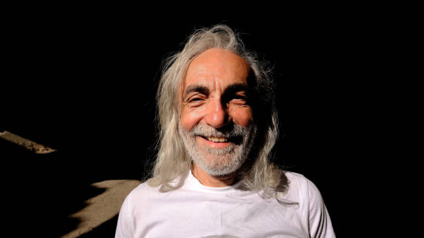 In a dark setting with a shadowy background, a beam of sunlight filters through, illuminating a 65-year-old man with white hair and a light beard. He stands in the light, wearing a playful and mischievous expression that brings warmth and character to the scene. The contrast between the darkness and the soft glow of sunlight on his face emphasizes his features, creating a visually striking portrait. This image effectively captures themes of warmth and personality within a moody environment.