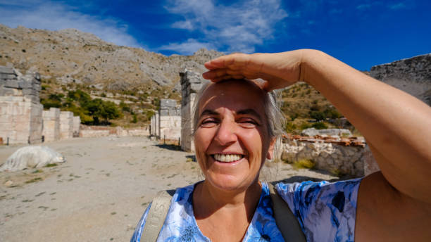 A wide-angle portrait of a Caucasian woman in her 60s with white hair and green eyes, carrying a backpack outdoors. She has a joyful and playful expression, adding a sense of adventure and positivity to the scene. The natural light enhances her features, while the blurred background provides depth, creating a vibrant and fresh atmosphere. Her cheerful demeanor, combined with the outdoor setting, makes this image versatile for stock photography, suitable for themes like travel, adventure, or enjoying nature in later years.