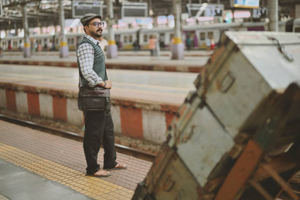 A mid-adult man stands on a railway platform, dressed in a stylish retro outfit that includes a cozy sweater and a sling bag, adding a touch of timeless charm. The sweater, wearing a glasses and the sling bag slung across his shoulder give him a distinctive, vintage appeal. His poised stance and thoughtful expression as he gaze the tracks reflect a blend of anticipation and nostalgia against the atmospheric backdrop of the railway station.
