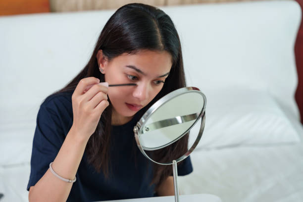 istock Asian woman applying mascara while sitting on bed, focused on enhancing eyelashes using a mirror for precision. Quiet bedroom environment with beauty tools laid out, part of morning makeup routine. 2178920075