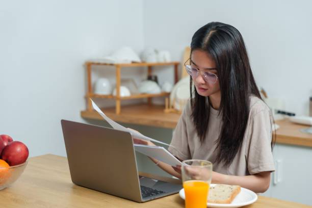 istock Asian woman working at kitchen table reviewing printed documents. Focused expression. Laptop, fruit bowl, and breakfast items placed on wooden counter. Multitasking during morning routine 2178920163