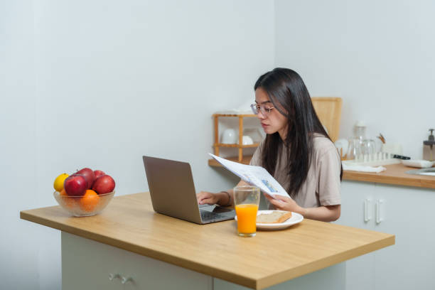 Young Asian woman working from kitchen table holding papers while using laptop. Bright indoor setting with food and fruit bowl on the side. Casual environment, multitasking with work and breakfast.