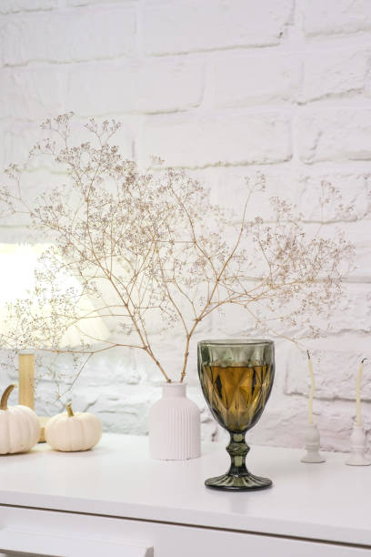 istock Home autumn composition.Charming green glassware with wine artfully combined with delicate decor of white pumpkins, lamp, dried branches on white bricks background. 2178921945