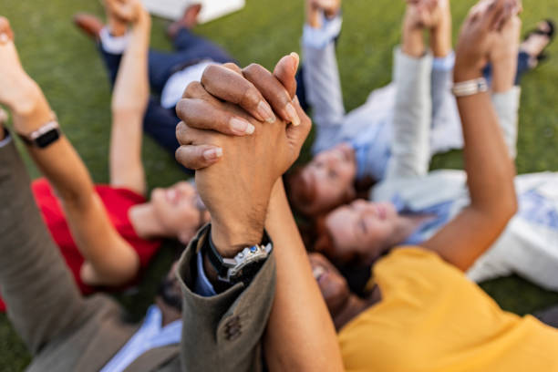 Diverse group of business people lying on grass, holding hands in a circle, showing unity and teamwork. Smiling faces reflect positive attitude towards work, teamwork, and success