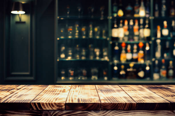 Real photographs material of rustic wooden bar countertop with a warm, ambient background of blurred bottles Real photographs material of rustic wooden bar countertop with a warm, ambient background of blurred bottles and shelves. pub table top view stock pictures, royalty-free photos & images