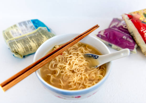 A bowl of instant noodles in soup base with a pair of chopsticks and ceramic spoon, and packets of the noodles isolated on white.