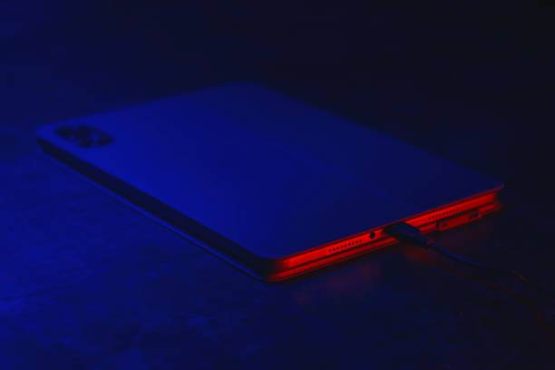 istock Digital tablet charges on the concrete table with a cable with technology lights, blue and red. Concept for charging the battery of gadgets. 2179146194
