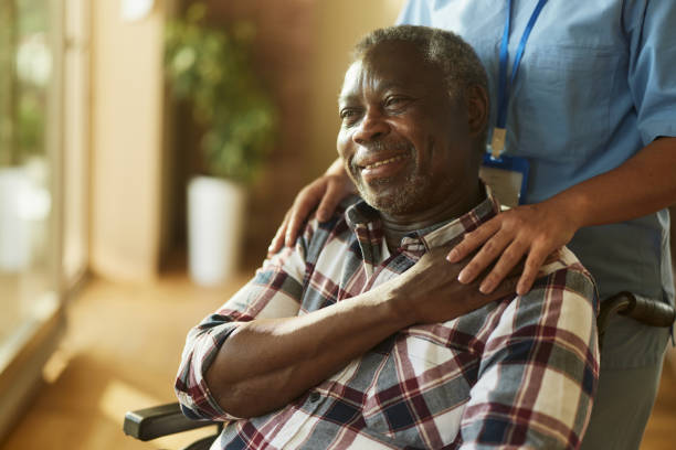 Happy black man in wheelchair having a support of his nurse. Happy African American mature man in wheelchair enjoying in support by his nurse at retirement home. sick black man stock pictures, royalty-free photos & images