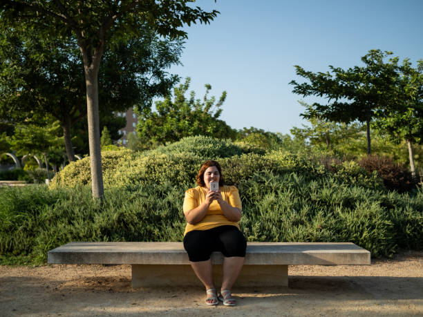 A woman with cognitive disability sits on a bench in a park, smiling as she engages with her smartphone. The sun shines brightly, illuminating the greenery around her.