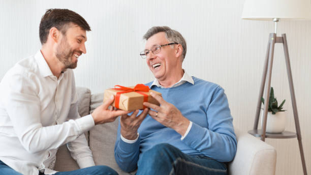 istock Son Congratulating Senior Father Giving Gift Sitting On Couch, Panorama 2179171310