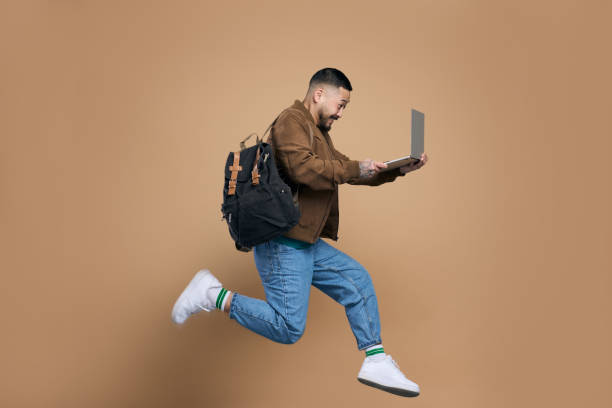 Excited student jumping with laptop and backpack on brown background
