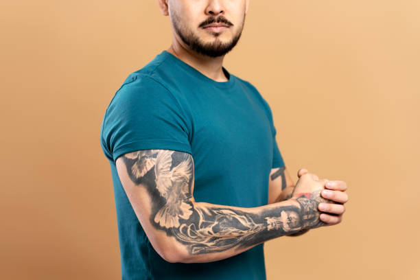Man with tattoos on his arms is standing with his arms folded in front of a beige background