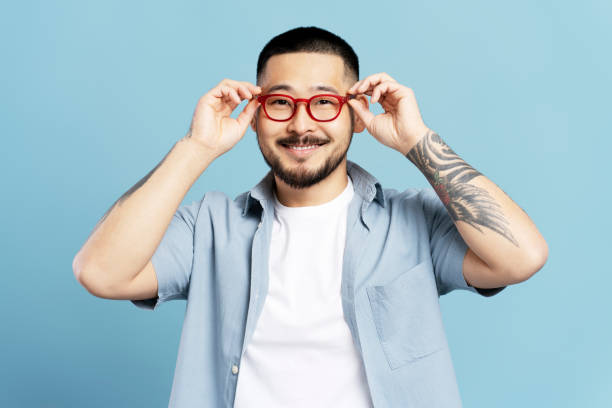 Young man with a beard and a tattoo is adjusting his eyeglasses and smiling on a blue background