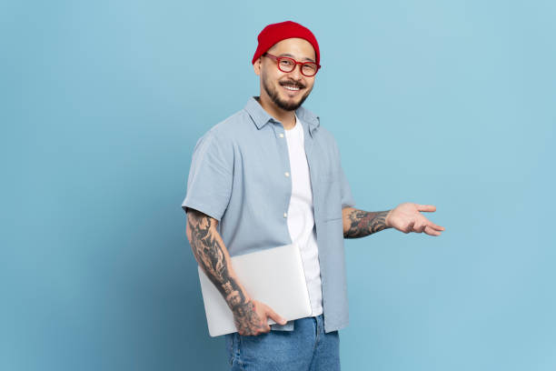istock Smiling asian man with tattoos holding laptop and gesturing with hand 2179188112
