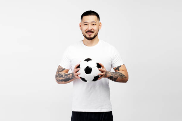 Young man with tattoos is holding a soccer ball and smiling on a white background