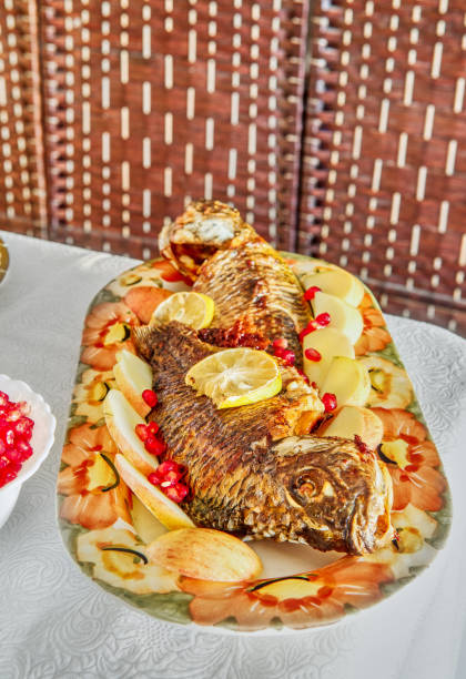 A beautifully roasted whole fish presented on a floral platter, garnished with fresh lemon slices and pomegranate seeds.