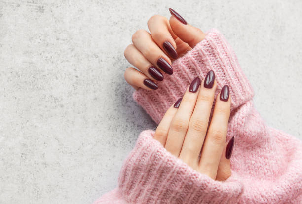Elegant maroon manicured nails displayed against a soft pink sweater on a textured gray background