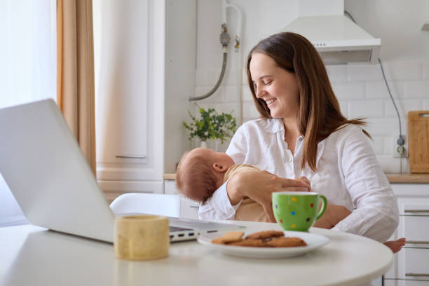 istock Woman smiling happily while working remotely from her kitchen on laptop with her newborn baby in hands doing her online job on her computer 2179223205