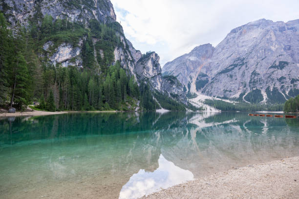 A serene alpine lake reflecting towering mountains and dense forests, with rocky shores and tranquil waters under a cloudy sky in a pristine natural setting.
