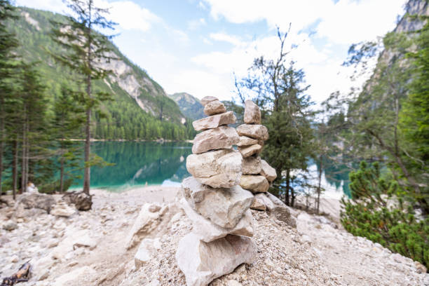 Carefully balanced rock cairns sit on a rocky shore, overlooking a calm alpine lake surrounded by forests and mountains in a peaceful natural setting.
