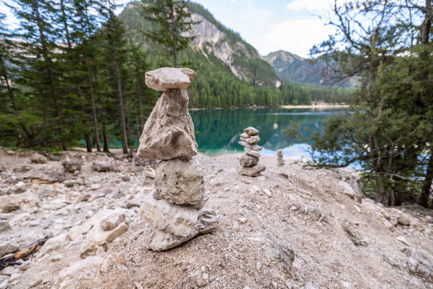 Carefully balanced rock cairns sit on a rocky shore, overlooking a calm alpine lake surrounded by forests and mountains in a peaceful natural setting.
