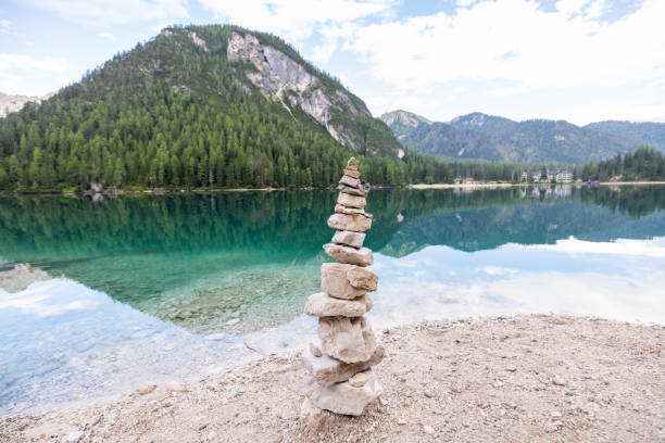 A balanced stone cairn stands by the shore of a clear alpine lake, reflecting the surrounding mountains and dense forest in a peaceful, natural setting.