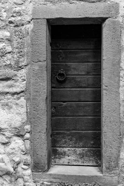 A black and white image of a wooden door set in a stone wall. The door is made of horizontal planks and has a simple iron ring handle. The stone wall is rough and textured.
