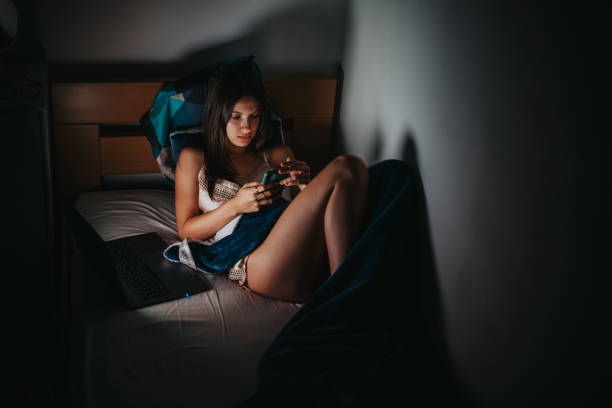A young woman comfortably sits in bed at night using her smartphone, illuminated by a warm glow. A laptop rests beside her, suggesting connectivity and relaxation.
