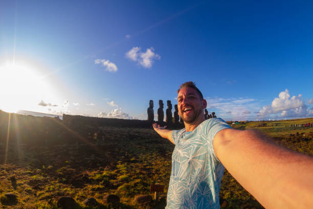A handsome young happiness cheerful joyful fit latin male backpacker tourist visiting Ahu Tongariki Easter Island - Rapa Nui, in the middle of Pacific Ocean, part of Polynesia / Chile in Latin America; posing for a picture in front of the Moai's sculptures with his open outstretched arms at Ahu Tongariki, wearing a blue t-shirt in a sunny summer sunrise moment. Man visiting and sightseeing Latin America destination. Latin America / Chile travel concept.