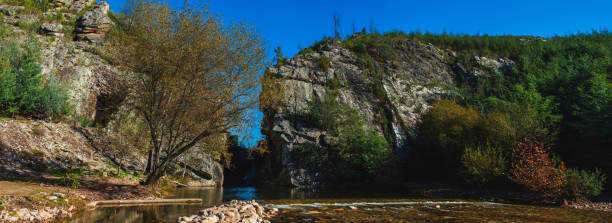 istock landscape showcases a river flowing between two large rock formations, with towering cliffs on either side. The cliffs are rugged and steep, featuring a mixture of stone and greenery, while trees and bushes grow along the slopes. 2179306346