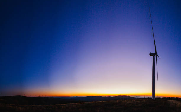 a mesmerizing sunset scene with wind turbines spread across a landscape. The sky transitions from a deep blue at the top, slowly fading into purple, and then into warm orange and yellow hues near the horizon
