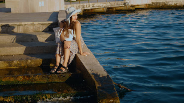 A woman is sitting on a ledge by the water. She is wearing a hat and a skirt