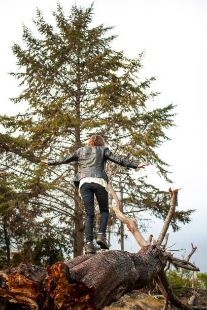 In a forest, a woman wearing a leather jacket and jeans is seen standing on a tree stump, blending fashion with nature. The scene conveys a sense of adventure and exploration. The setting is natural, surrounded by tall trees and a peaceful atmosphere.