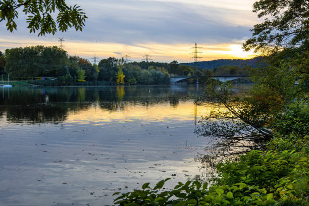 River Neckar in Heidelberg in summer sunset seen from bridge. View downstream to western outskirts of city. A few people are in street. Buildings are on both sides of river