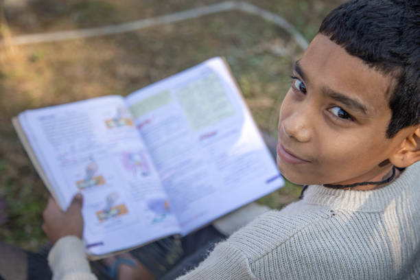 Poor Indian boy sits comfortably in a chair, engrossed in a book, surrounded by greenery. Bright sunlight filters through the leaves, creating a peaceful atmosphere.