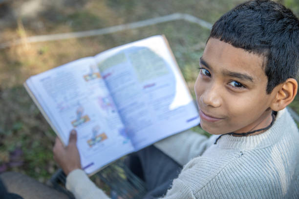 Poor Indian boy sits comfortably in a chair, engrossed in a book, surrounded by greenery. Bright sunlight filters through the leaves, creating a peaceful atmosphere.
