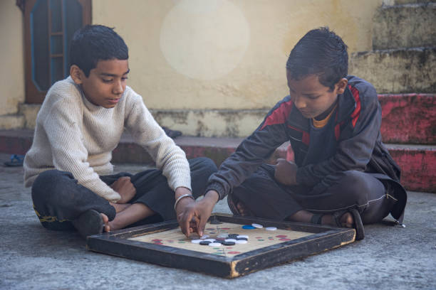 Two Indian village children are playing a carrom board game outdoors on a concrete surface. The children are sitting cross-legged, focused on the game.