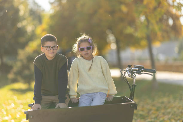 Portrait of brother and little sister sitting in cargo bike and relaxing in sunny autumn park